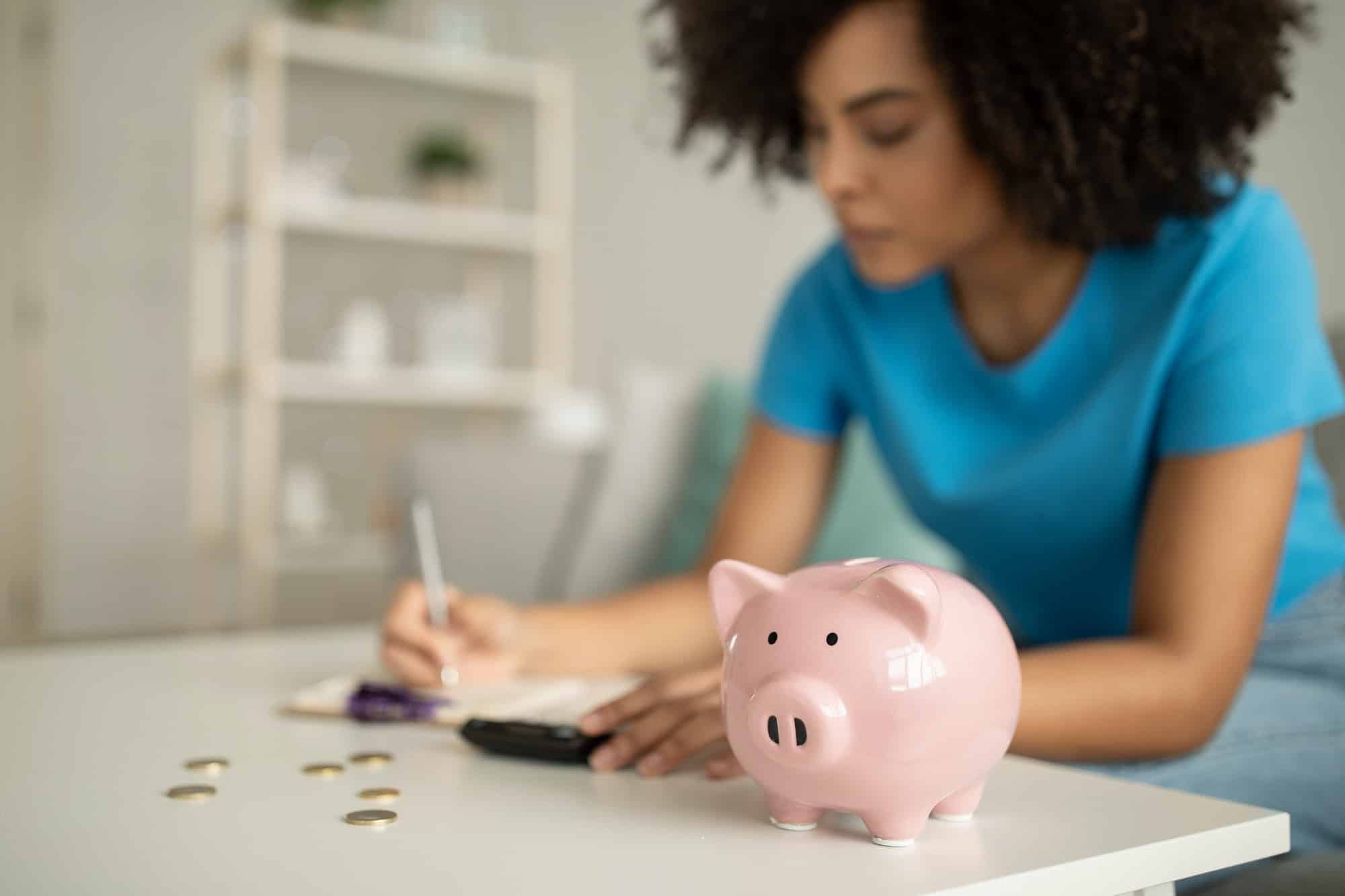 Serious concentrated millennial african american curly lady counting money near piggy bank in living