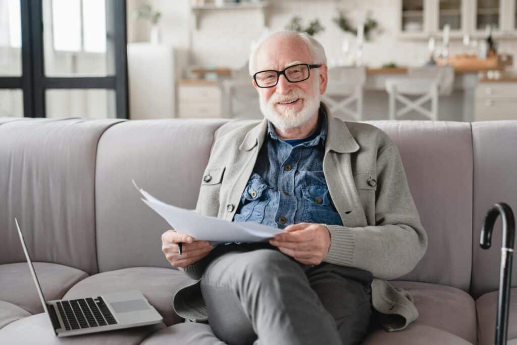 Grandfather looking at camera checking pension documents, loan, e-banking, paperwork with laptop